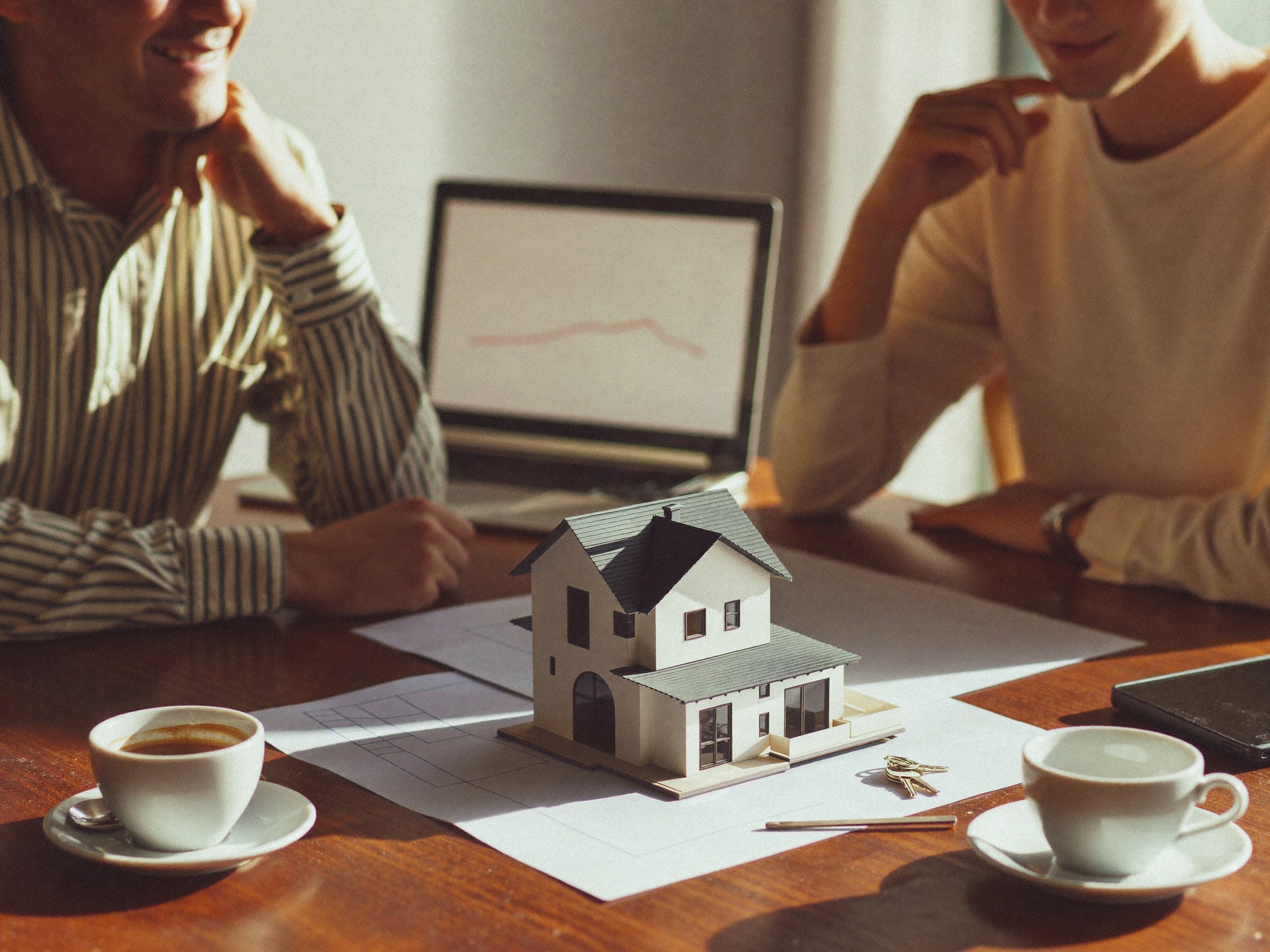 An investor and a property owner reviewing plans and a house model together at a sunlit table.